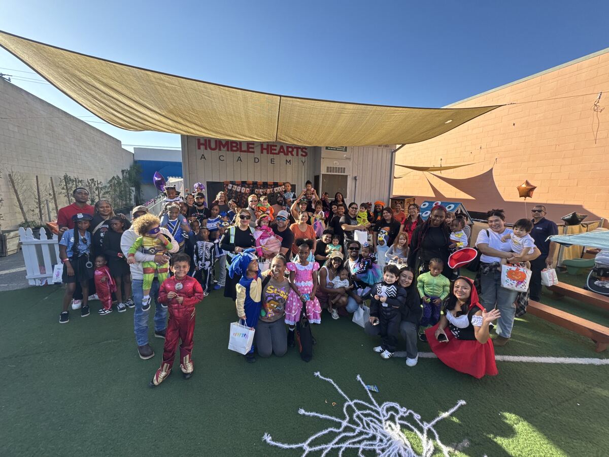 Children, families and staff posing together in the outdoor yard in front of the Humble Hearts Academy sign