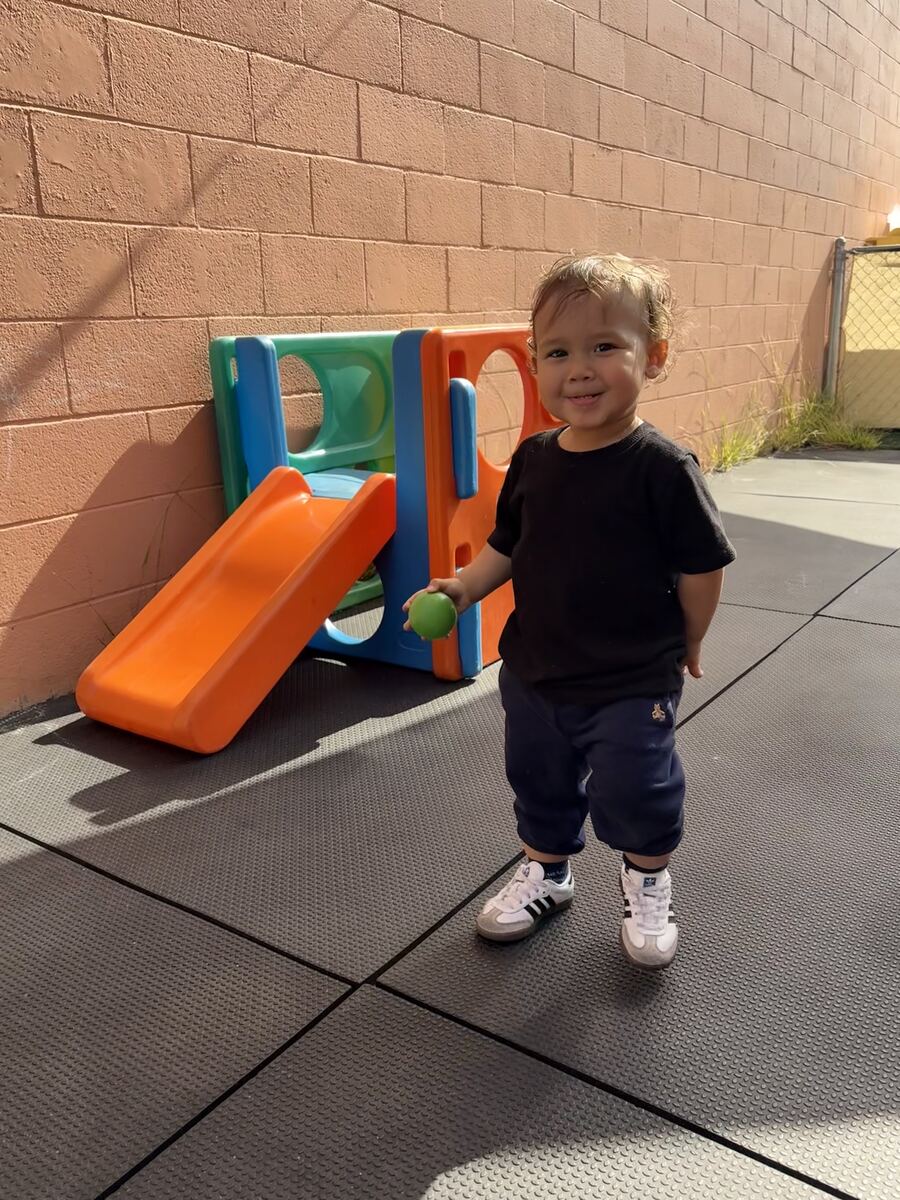 Smiling toddler holding a ball beside a small slide in the outdoor play yard