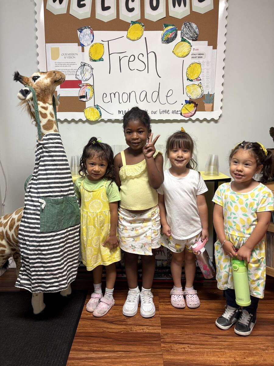 Four smiling preschoolers in lemon-print outfits posing in front of a welcome board in the classroom
