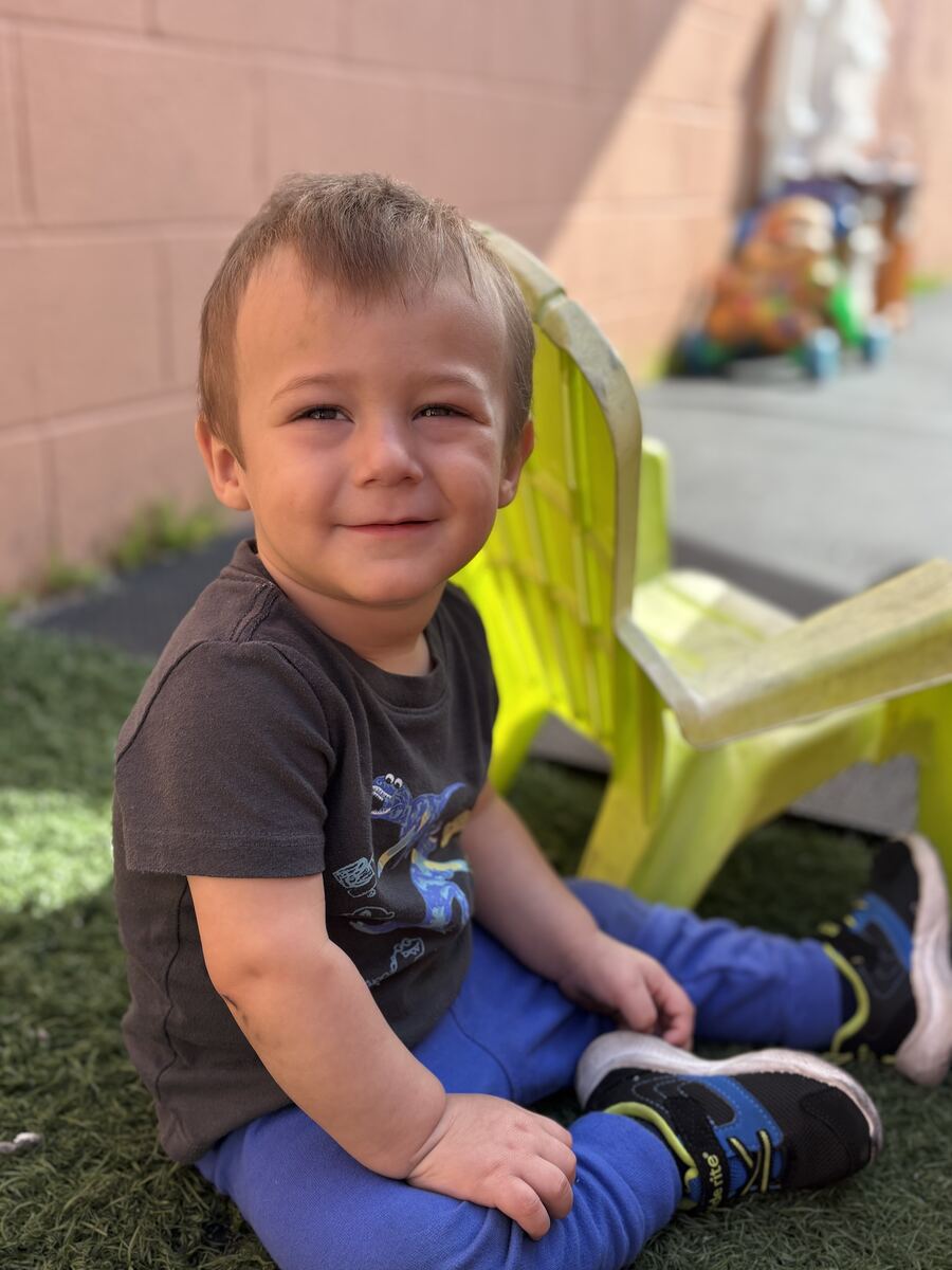 Smiling toddler sitting on the turf next to a yellow outdoor chair