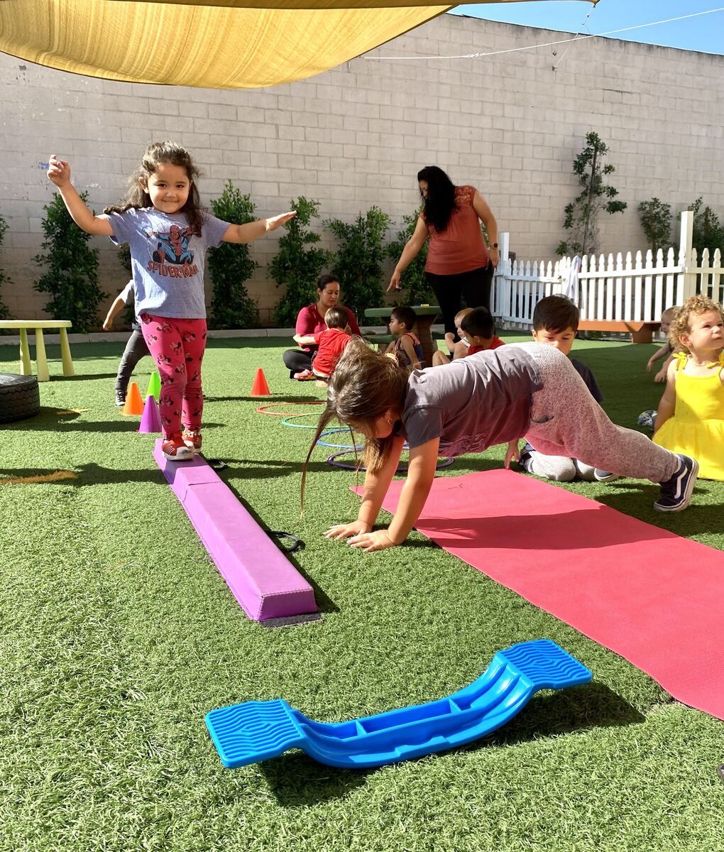 Preschooler walking across a purple balance beam on the playground while teachers and classmates play nearby